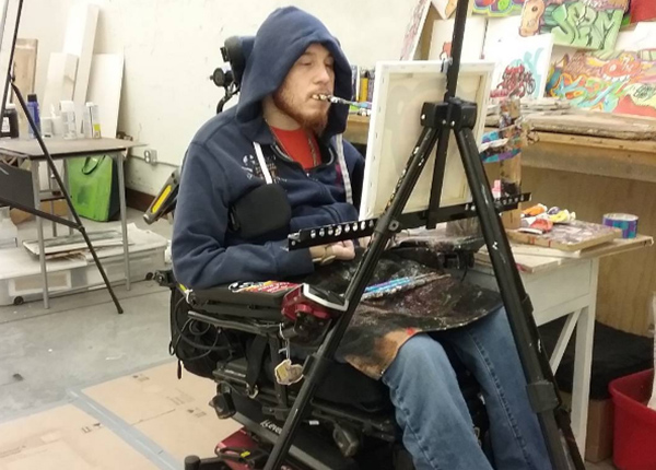 Man in wheelchair paints holding a brush in his mouth