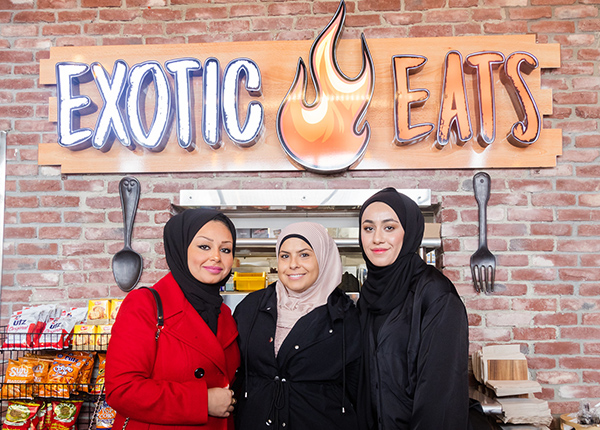 Three women wearing hijabs stand in front of a sign that says Exotic Eats