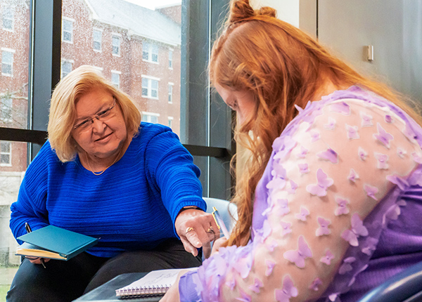 Two women sitting together, one of them points at a notebook the other has in her lap