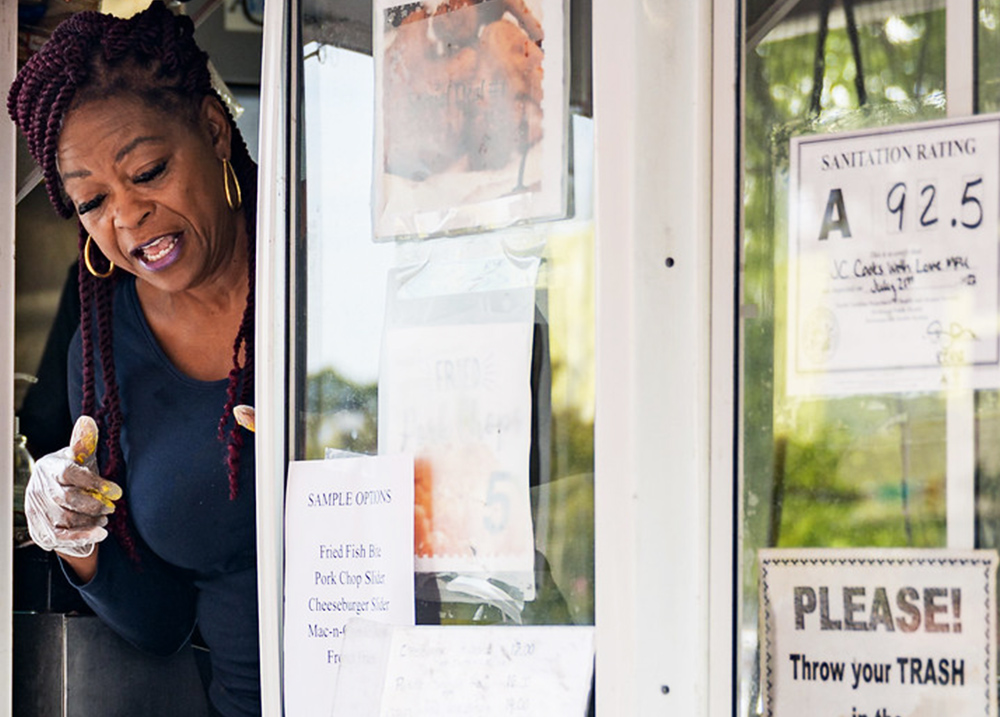 Woman talking to someone out of the window of a food truck that has a sign with a sanitation rating of 92.5