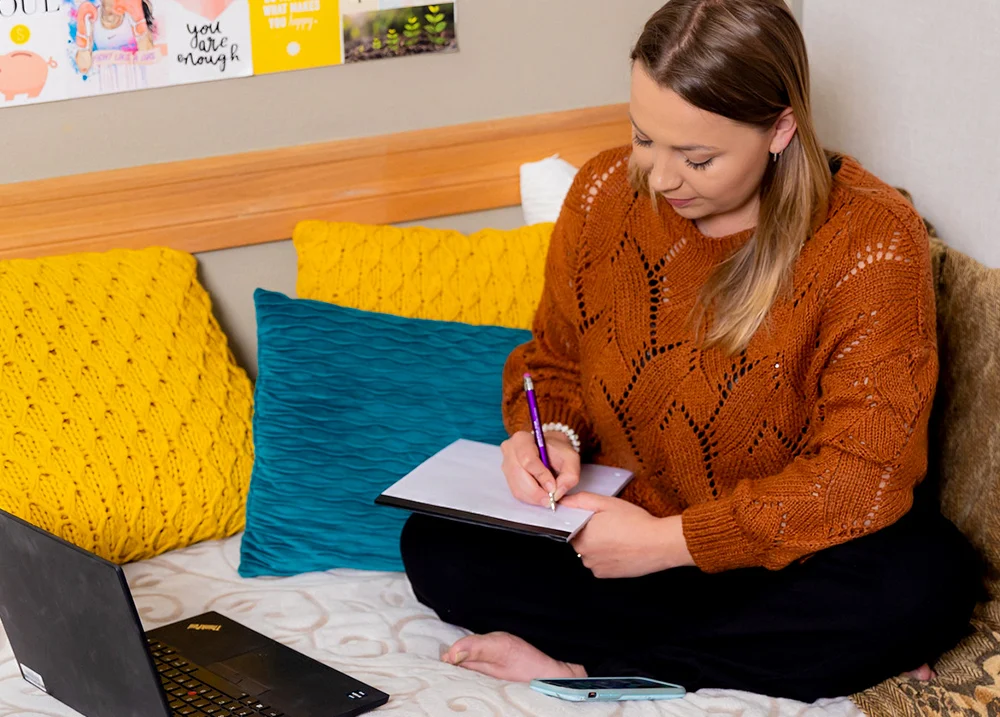 Woman sitting writing in a notebook with a laptop in front of her