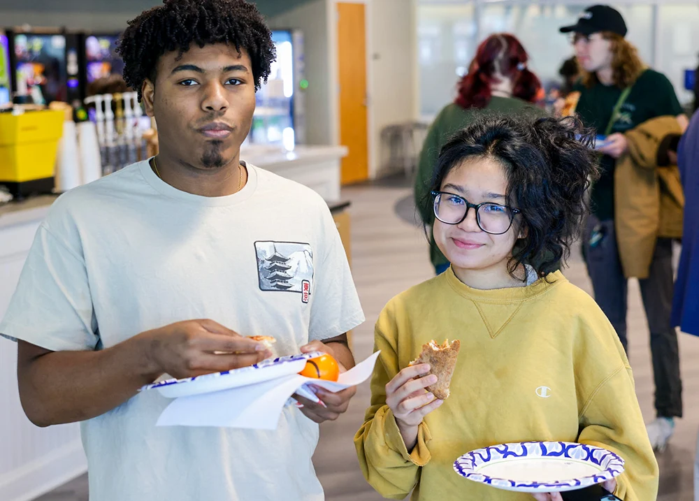 Two students standing next to each other eating pizza