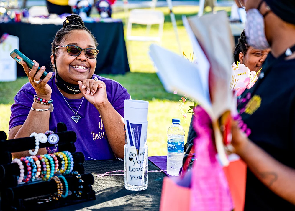 Woman sitting at an outdoor vendor table smiling as a customer approaches