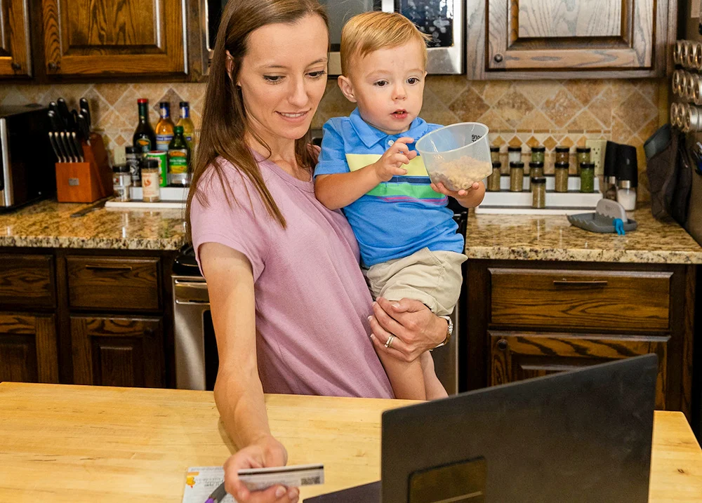 Mother holding her son in one arm while she looks at a laptop and holds a credit card in the other hand