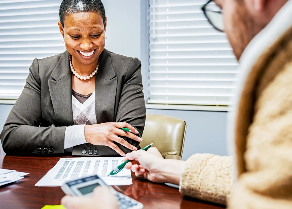 Two people sitting at a desk, one holds a calculator and points to a printed spreadsheet