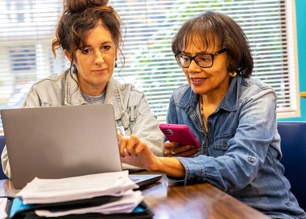 Two women sitting at a laptop, one holds a phone and points to something on the screen