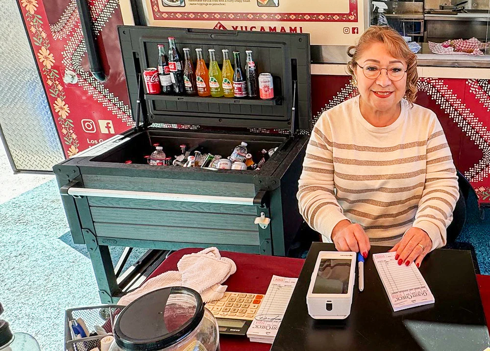 Woman sitting at a table in front of a food truck with a payment terminal and order pad in front of her